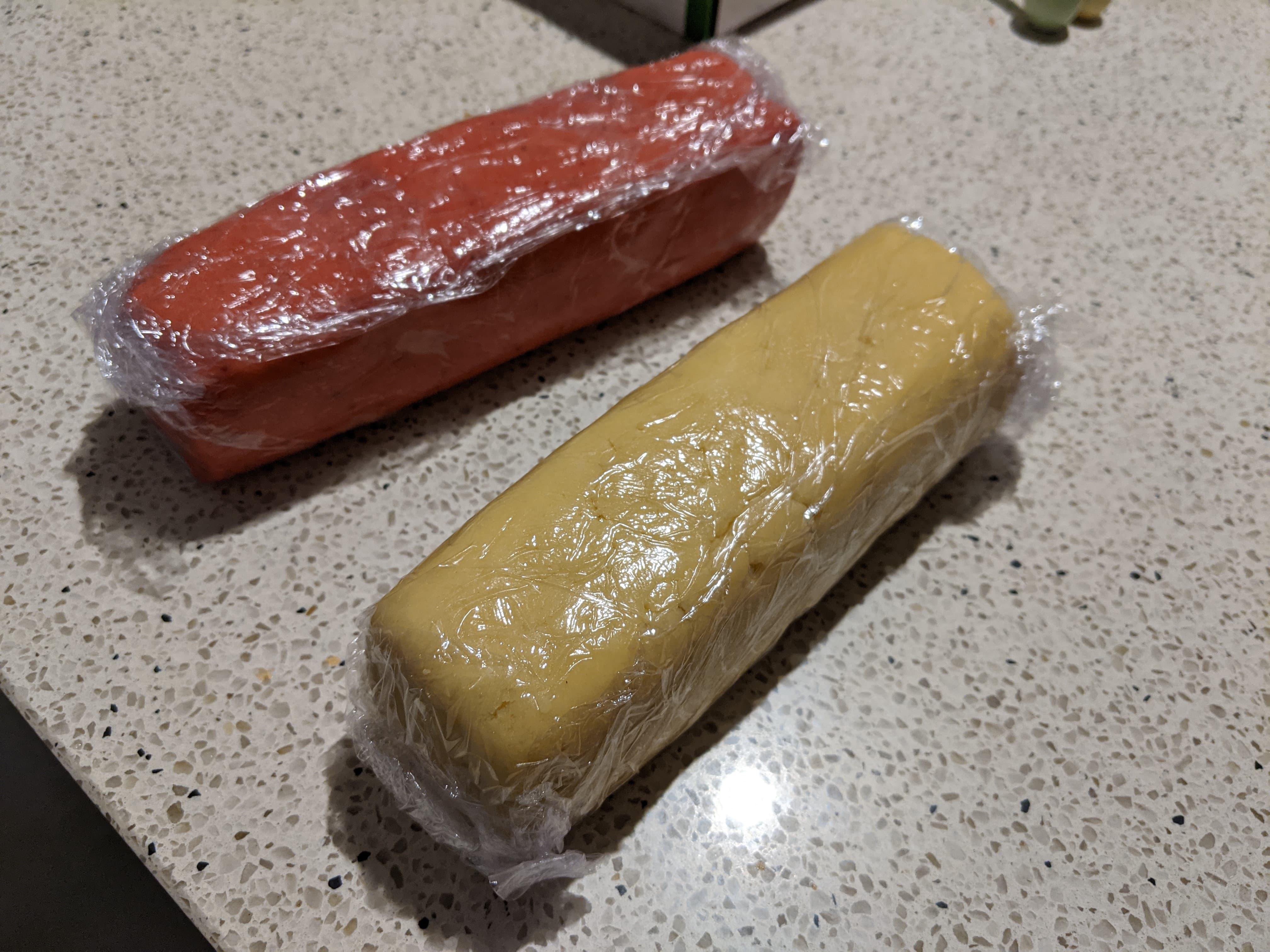 Two separate square-prism shaped logs of cookie dough wrapped in plastic, one red and one white