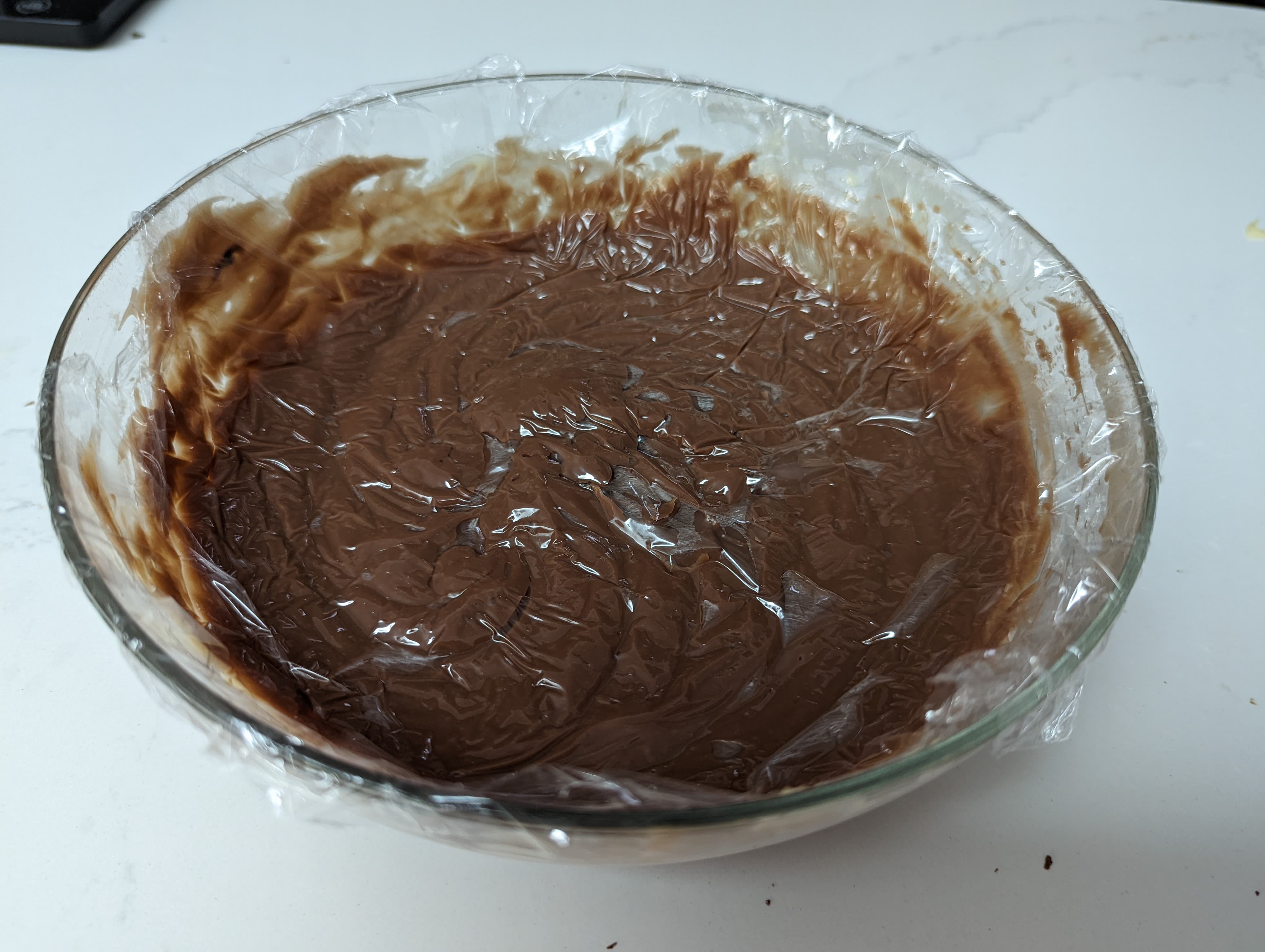A glass bowl filled with chocolate cream, with plastic wrap pressed to the surface of the cream